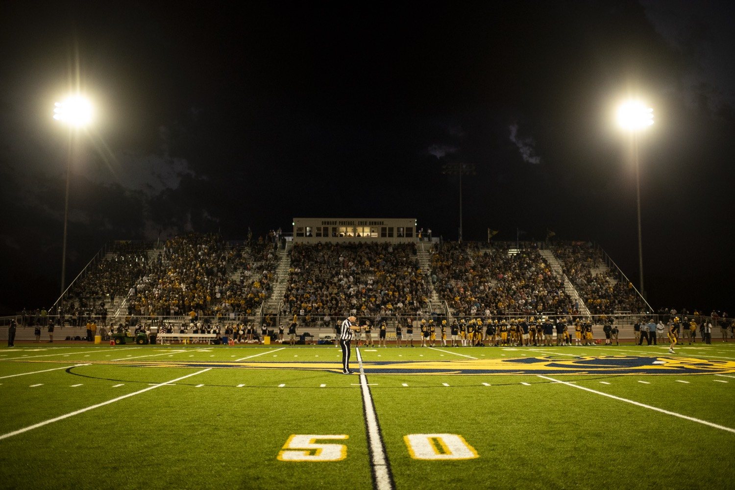 A high school football field at night, the 50-yard line in front of packed yellow stands